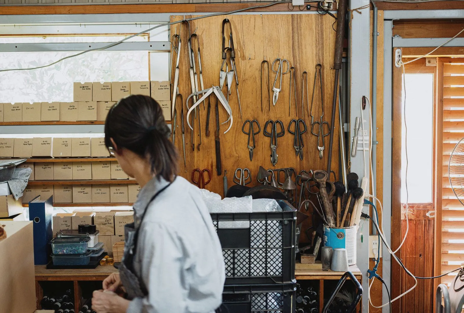 A glass artisan in a Nagano workshop, with tongs and shears hanging on a wooden wall and boxed stock on shelves