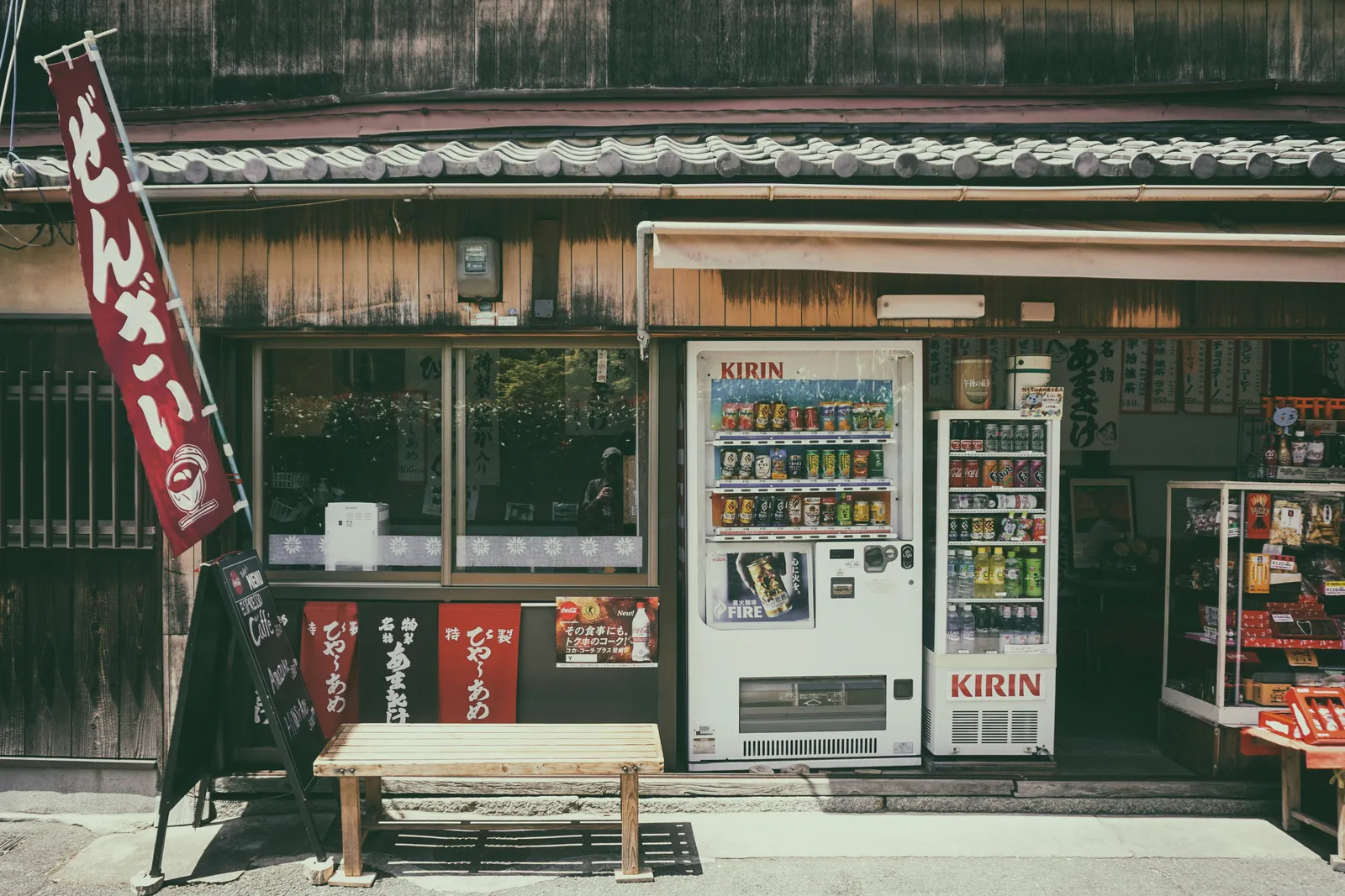An old wooden shopfront in Japan with two Kirin drink vending machines, a wooden bench and a hanging banner