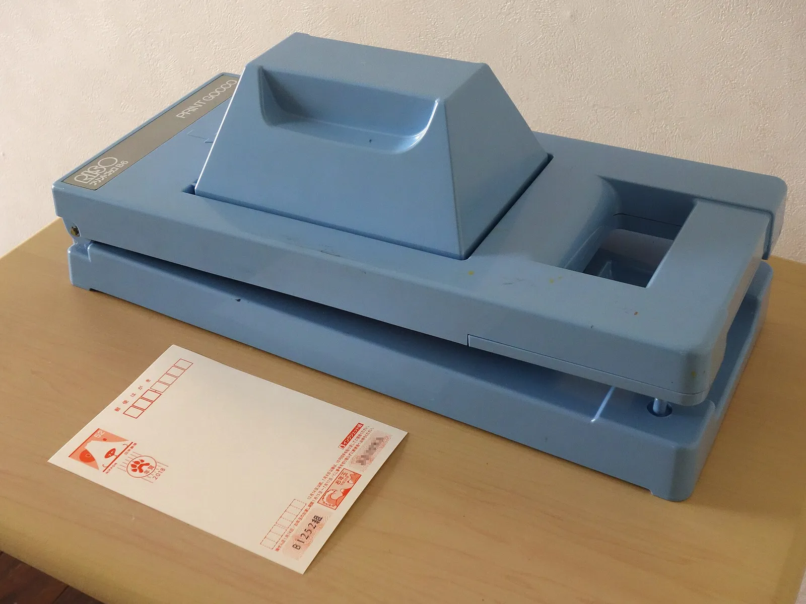 A blue Print Gocco home screen printer on a table beside a blank Japanese New Year postcard