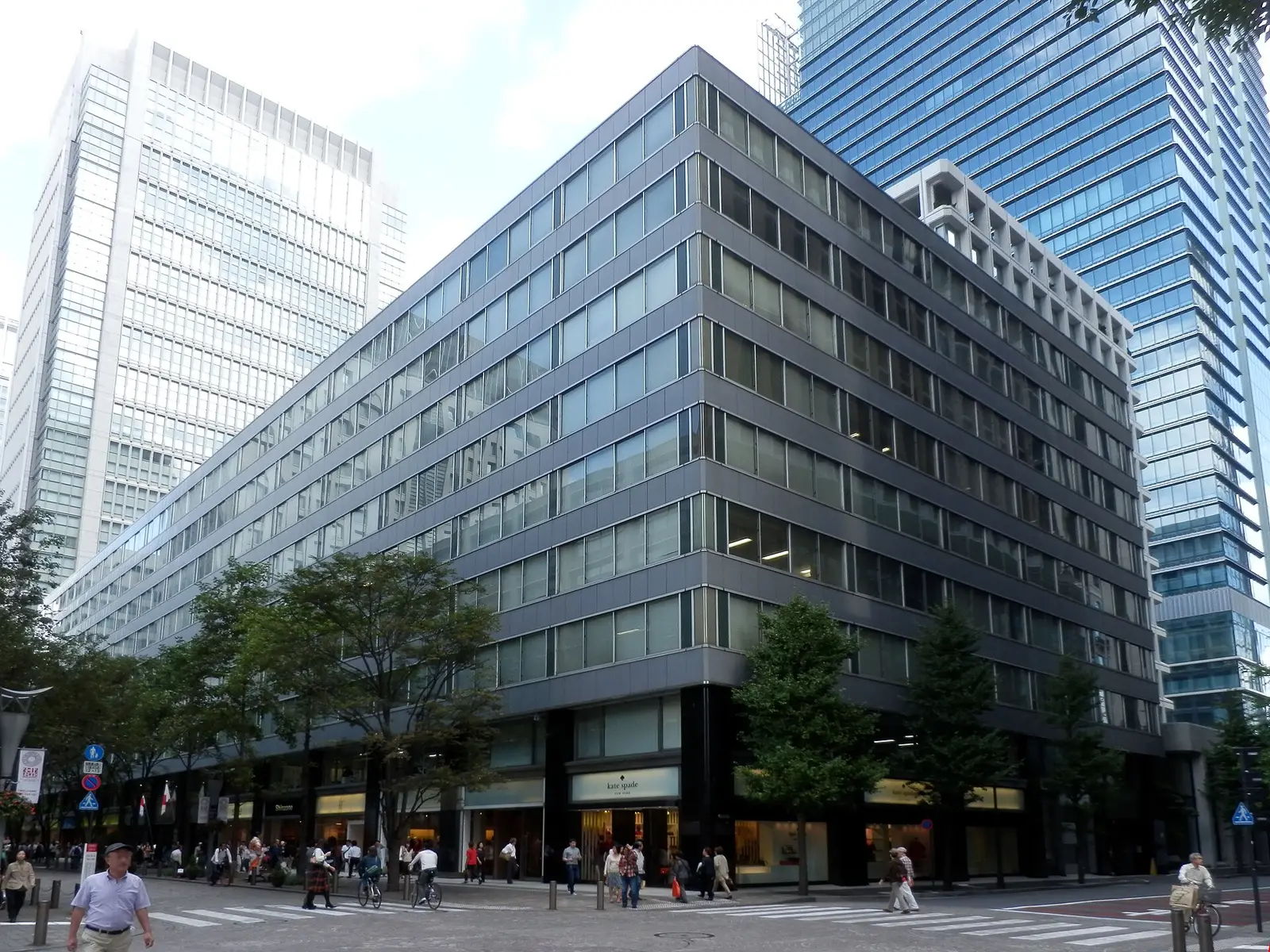 Glass and stone office buildings on a Marunouchi street corner in Tokyo, with pedestrians crossing below