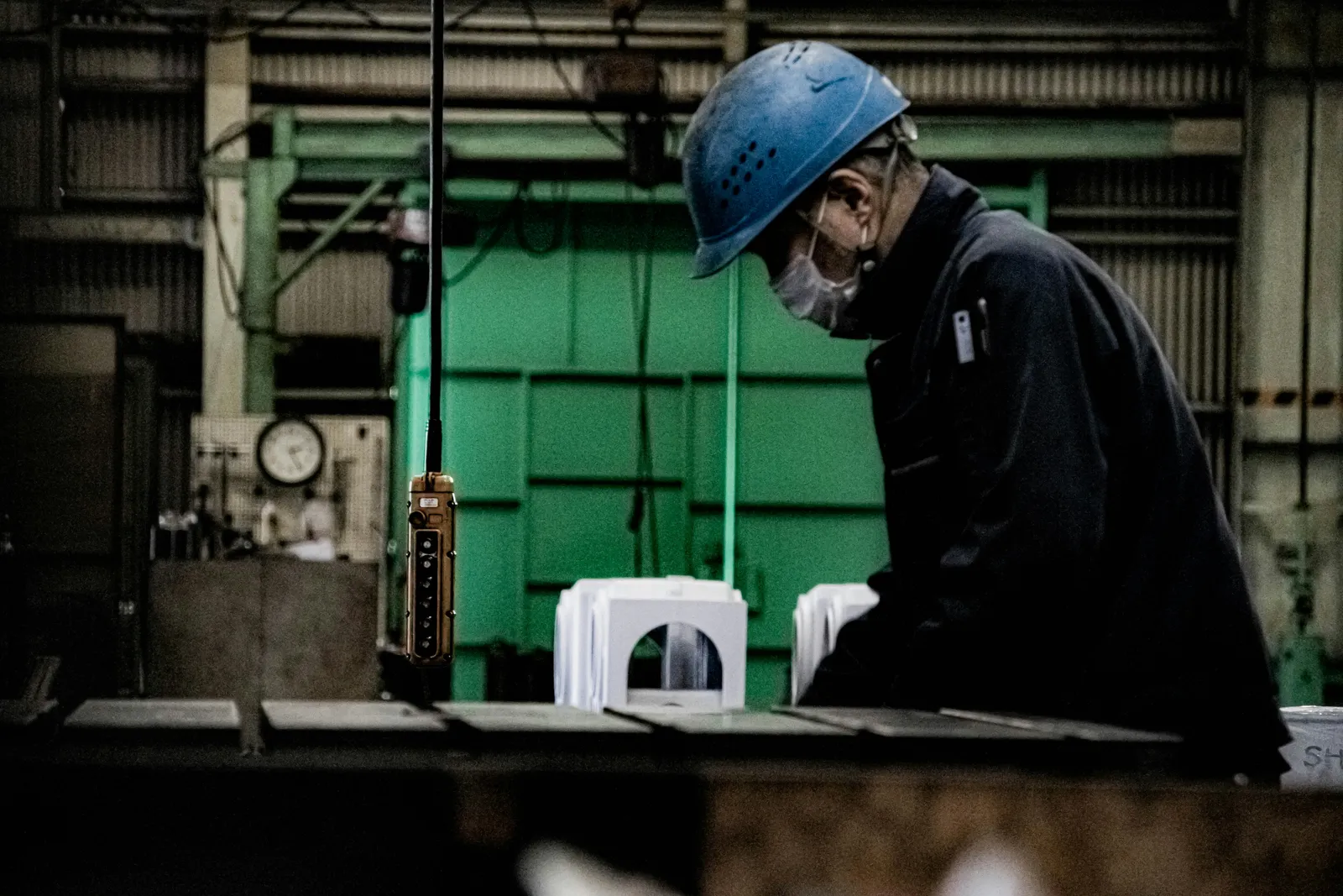 A worker in a blue hard hat and face mask bending over a workbench inside a factory