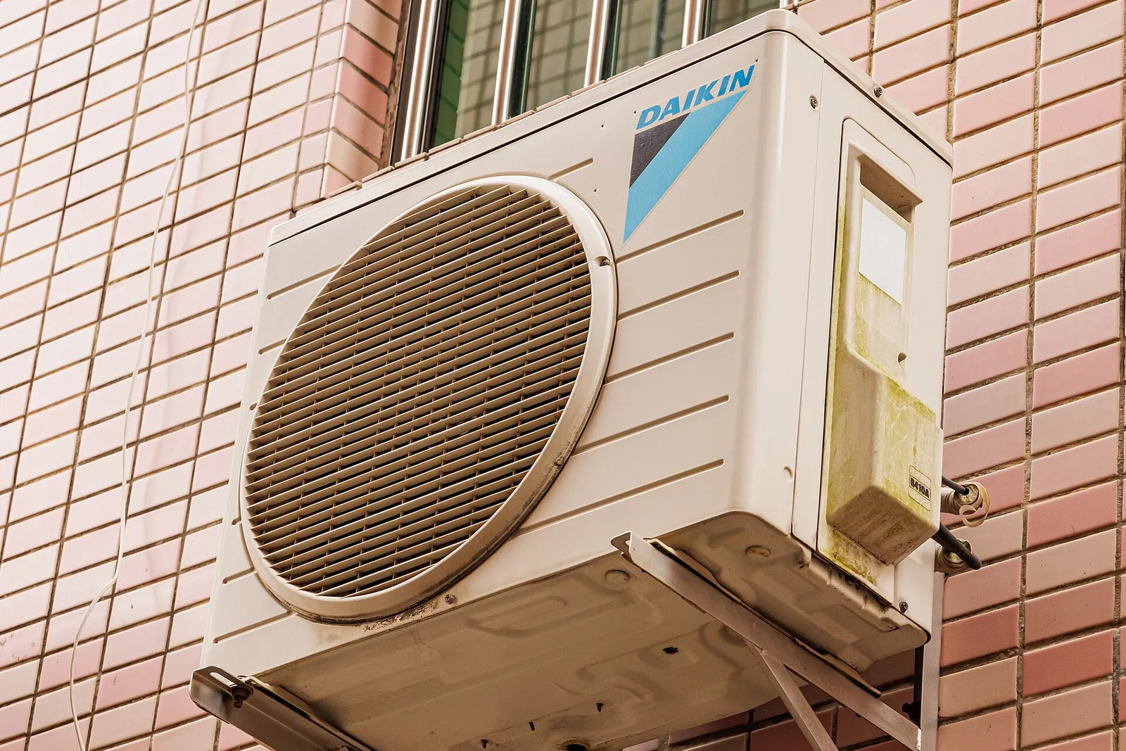 A Daikin air conditioner outdoor unit mounted on a metal bracket against a pink tiled building wall