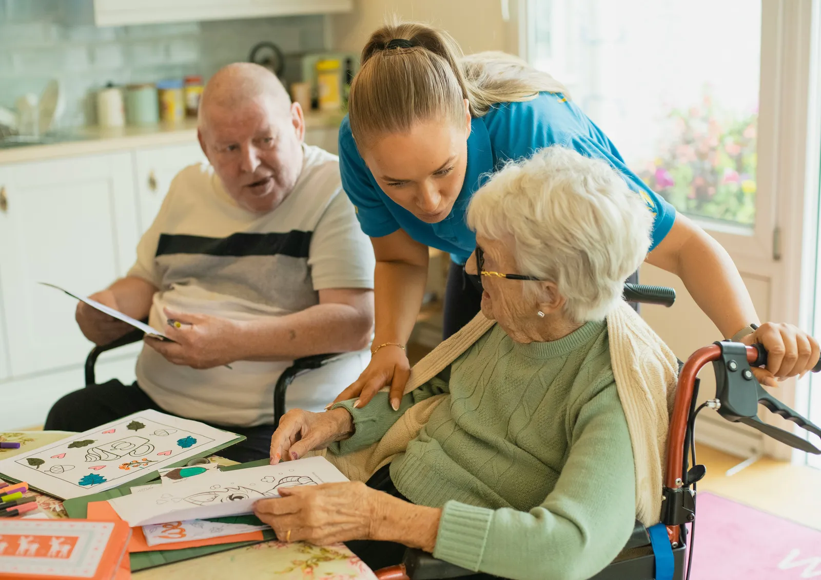A caregiver in a blue uniform helping an older man and a woman in a wheelchair with a coloring activity