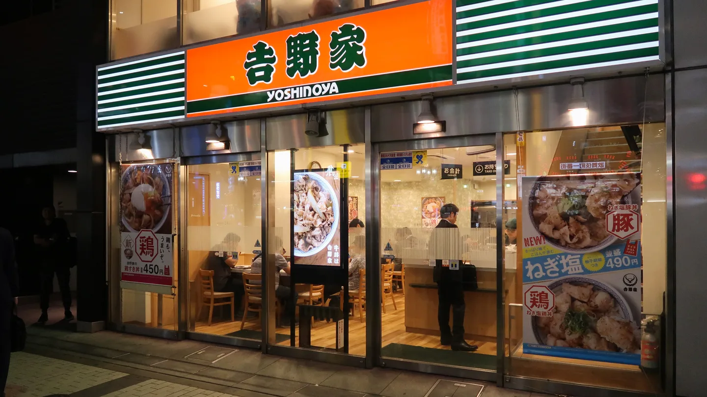 The glass front of a Yoshinoya restaurant in Shinjuku at night, with the orange 吉野家 sign lit above the door and menu posters in the windows