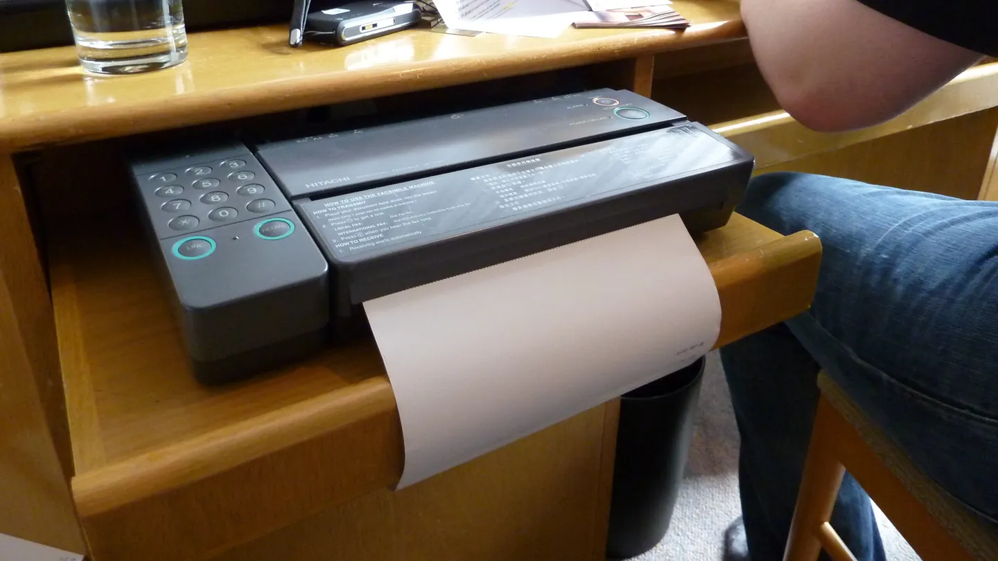 A compact fax machine in a desk drawer printing a page while a person types on a laptop beside it