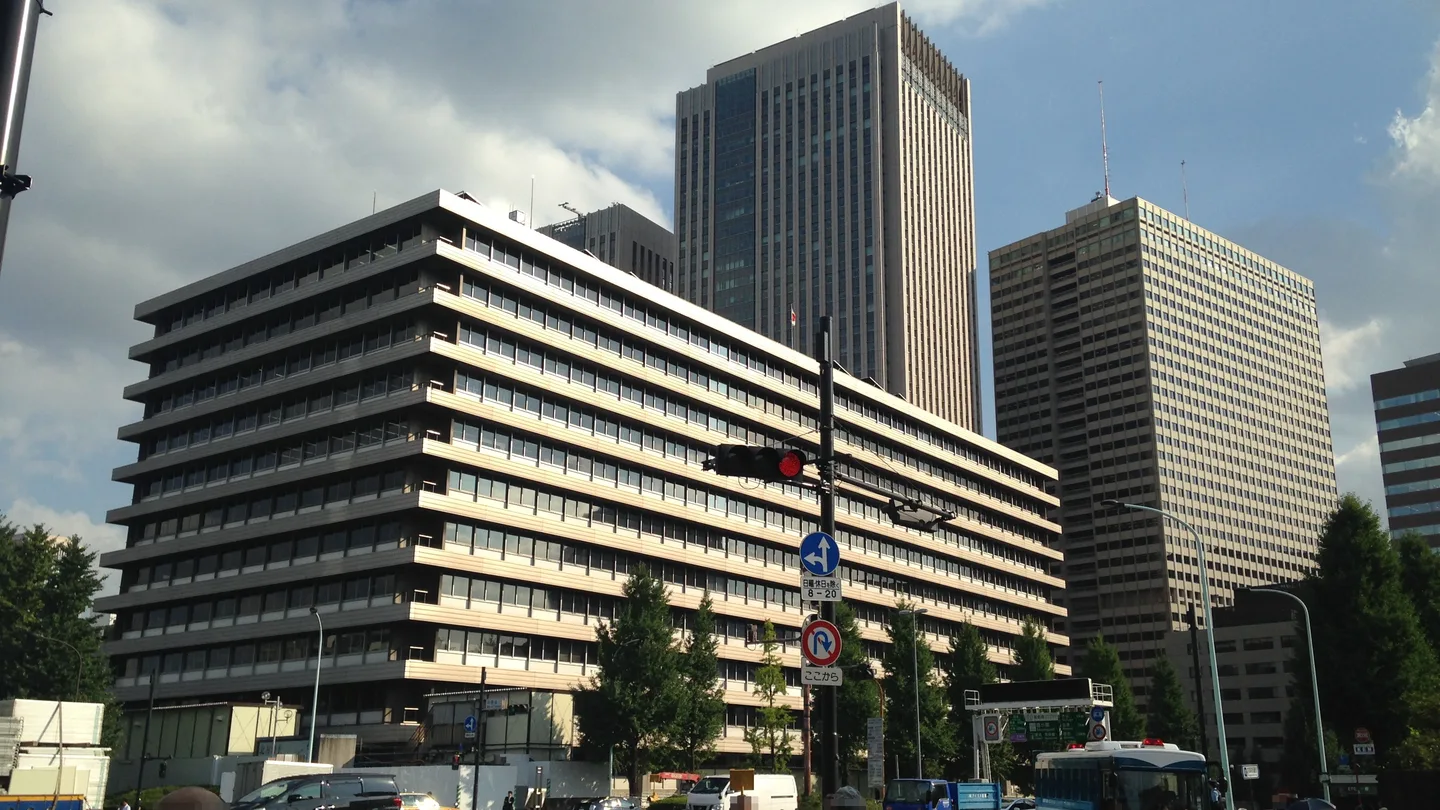 Two government office blocks in Kasumigaseki, Tokyo, a long horizontal building in front and a tall tower behind, with a traffic light and road signs in the foreground under a cloudy sky