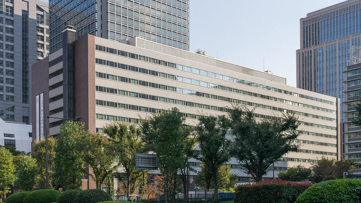 A long, pale, ten-storey government office building in Kasumigaseki behind a row of green trees, with glass towers rising behind it