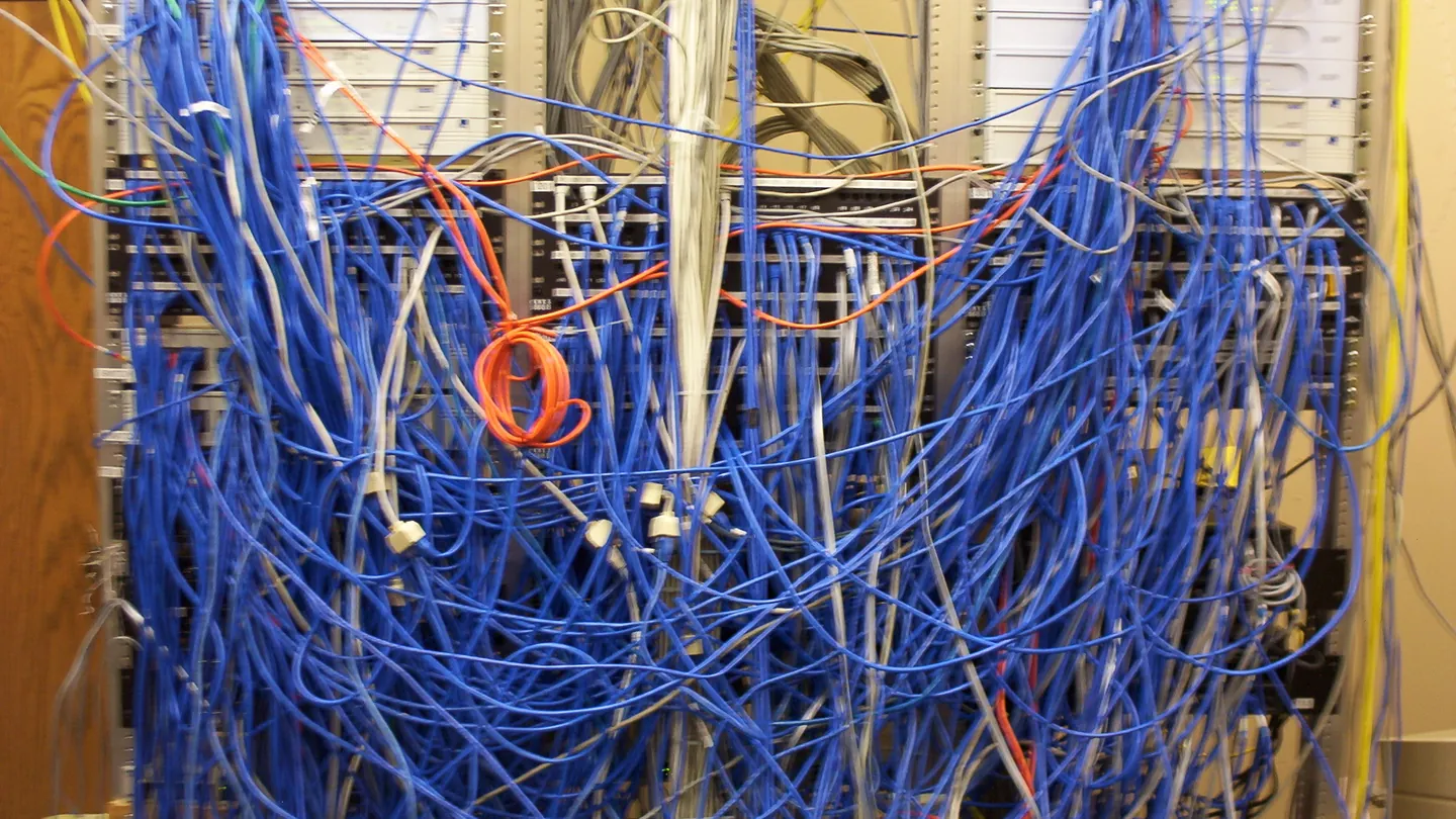 A wall-mounted network rack buried under a dense tangle of blue, orange, and yellow cables