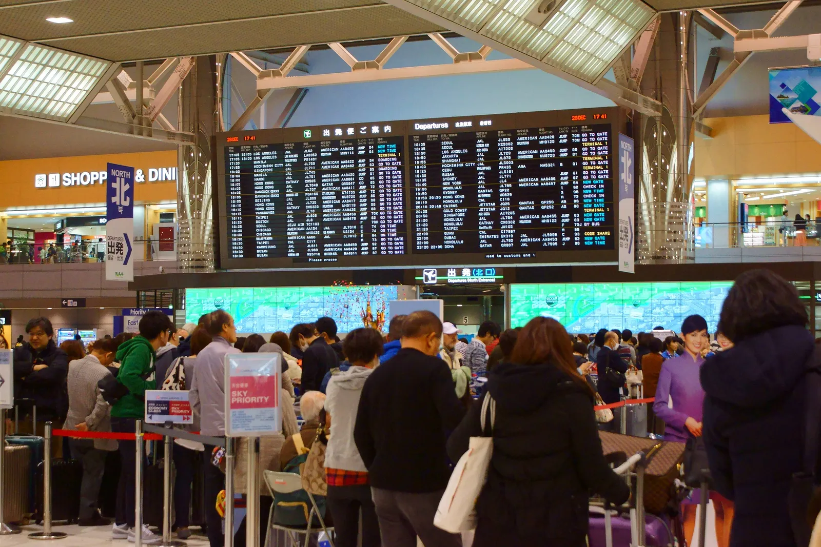 Travelers with luggage queueing at check-in counters beneath the departures board in Narita Airport Terminal 2