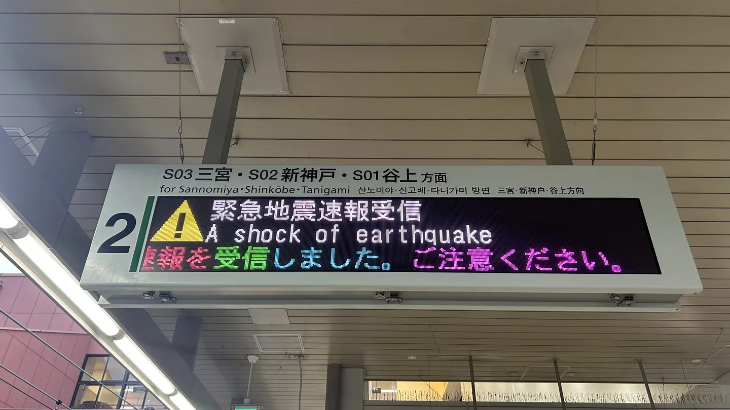 A platform departure display at a Kobe subway station showing an Earthquake Early Warning in Japanese and English, with a yellow warning triangle, hanging from a station ceiling