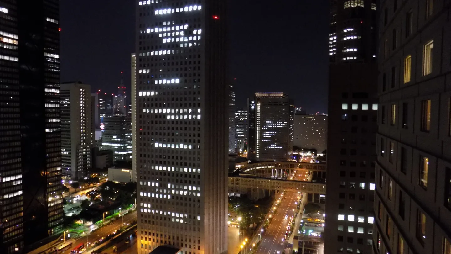 Nishi-Shinjuku office towers at night, their lit windows visible from a neighboring building, with traffic on the streets below