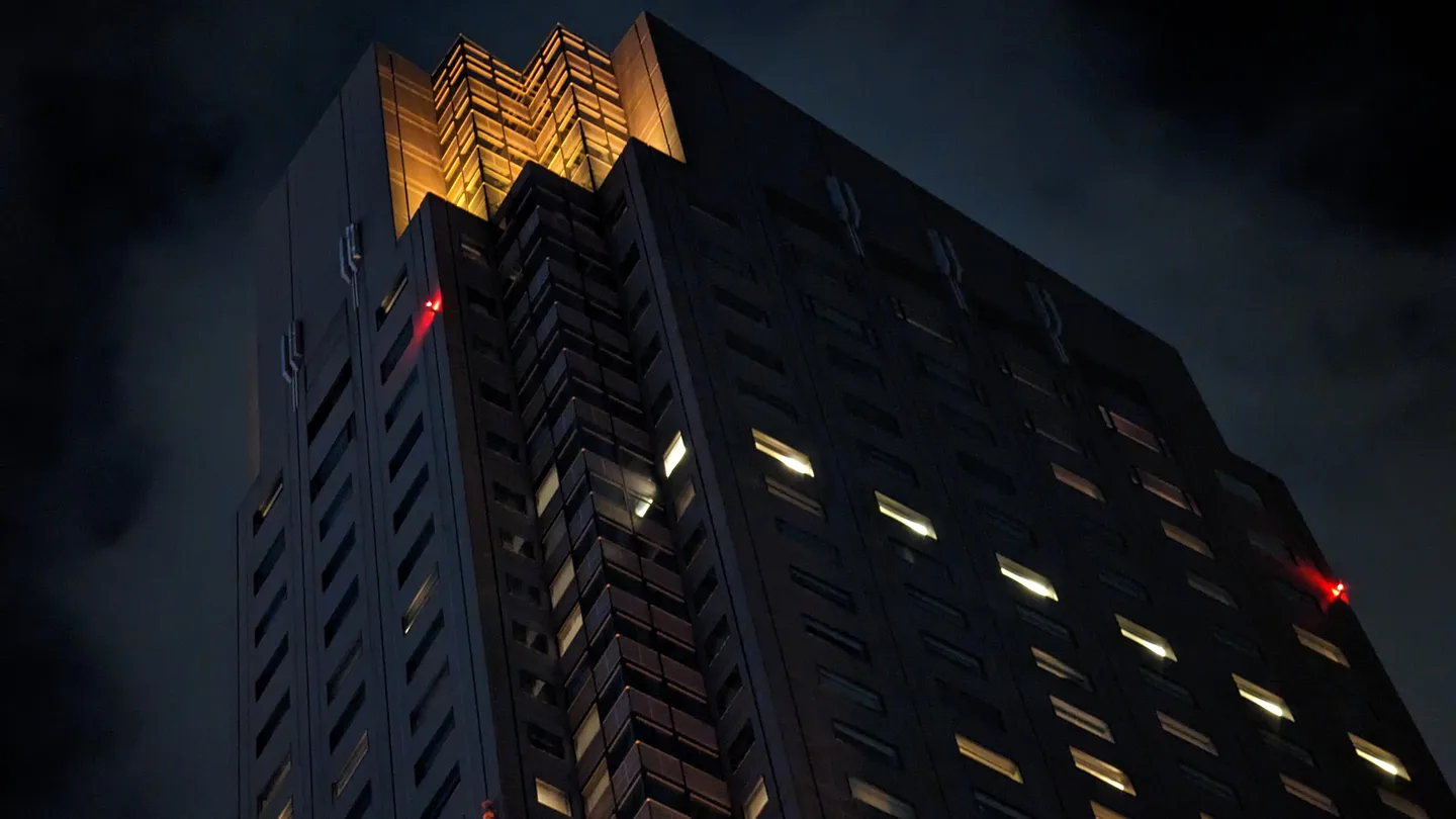 The top of a dark, angular skyscraper at night, its crown lit gold and a few office windows glowing, against a cloudy night sky