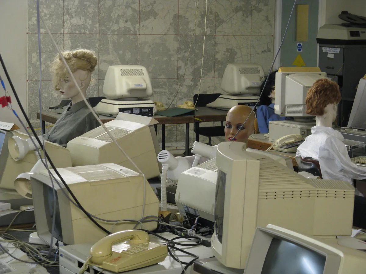 Beige CRT computers and desk phones crowded into a disused office, with mannequin heads seated at the desks