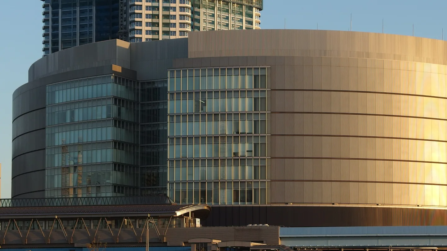 A curved, metal-clad data center building in Toyosu, Tokyo, lit gold by low winter sun, with glass stair towers, residential high-rises behind it, and an elevated walkway in front