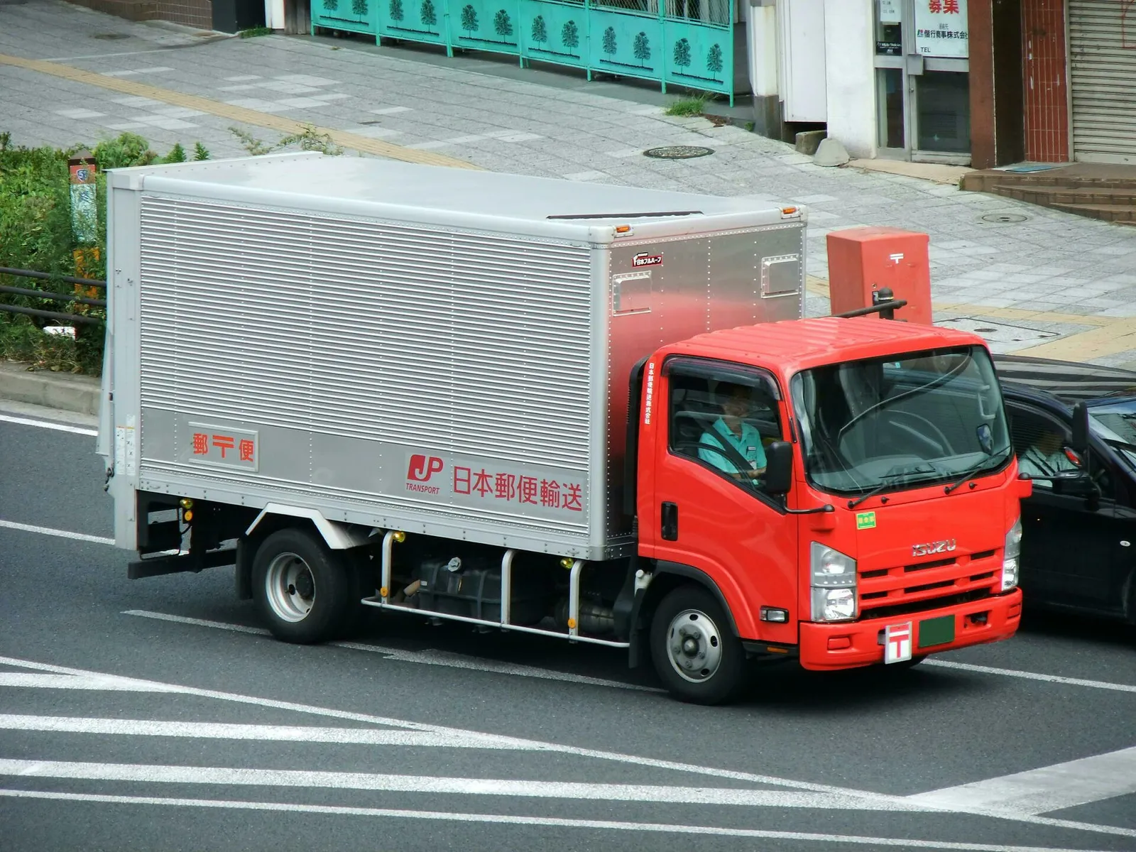 A red Isuzu Elf truck with a silver cargo box marked Japan Post Transport driving on a city street