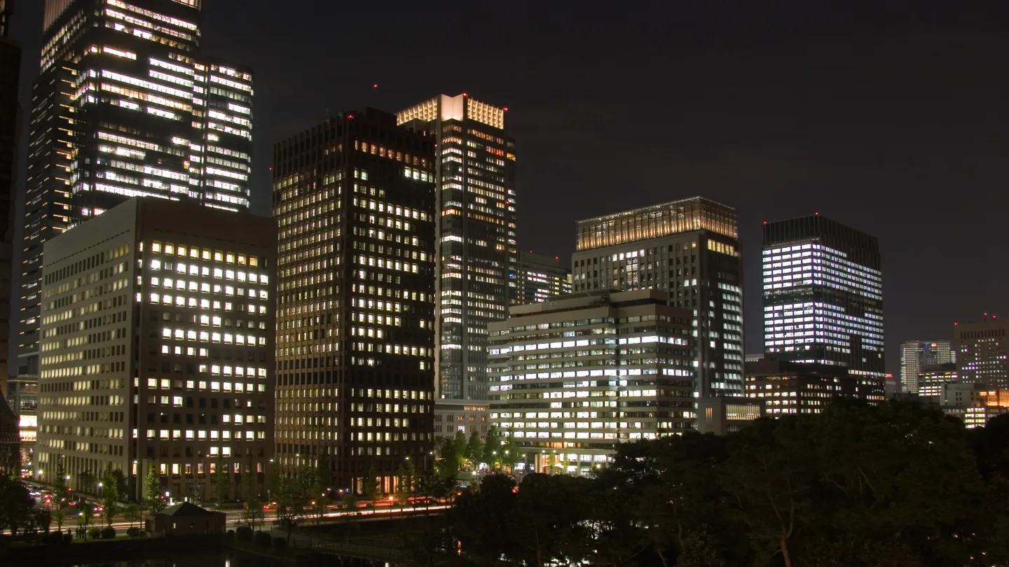 Office towers in Marunouchi, Tokyo, at night, with rows of lit windows visible across the trees of the Imperial Palace moat