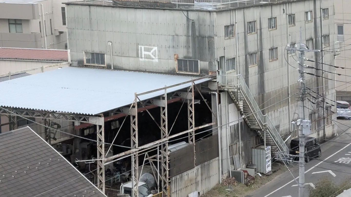 A weathered multi-storey factory building with an open steel-framed work shed beside a narrow street