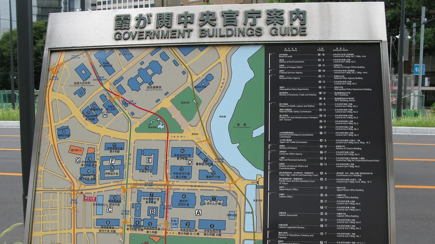 A street sign titled 霞が関中央官庁案内, Government Buildings Guide, showing a colour map of the ministry buildings beside the Imperial Palace moat, with a list of ministries and agencies in Japanese and English