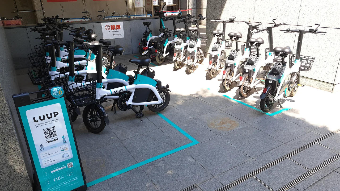 A row of white and teal Luup electric scooters parked in a marked bay beside an office building in Marunouchi, Tokyo, with a Luup sign and QR code in the foreground