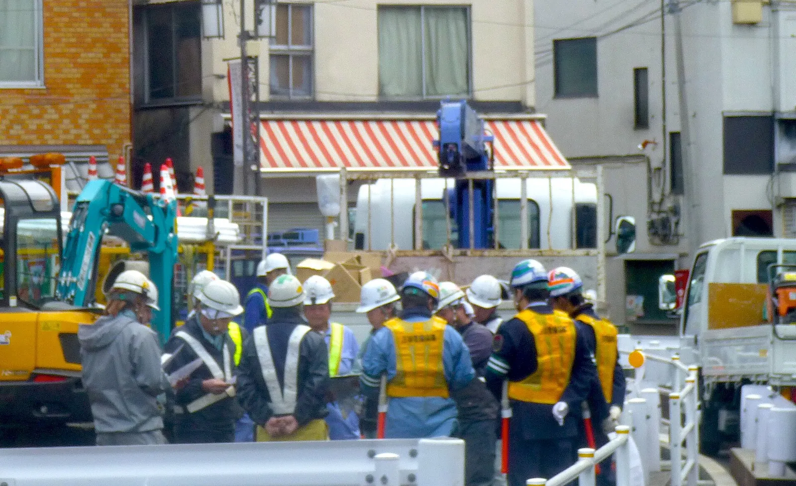 Construction workers in hard hats and safety vests gathered beside an excavator at a street work site
