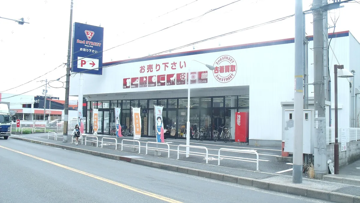 The white front of a 2nd STREET secondhand store beside a road in Higashi-Osaka, with its blue roadside sign, red banner of goods it buys, and bicycles parked by the entrance