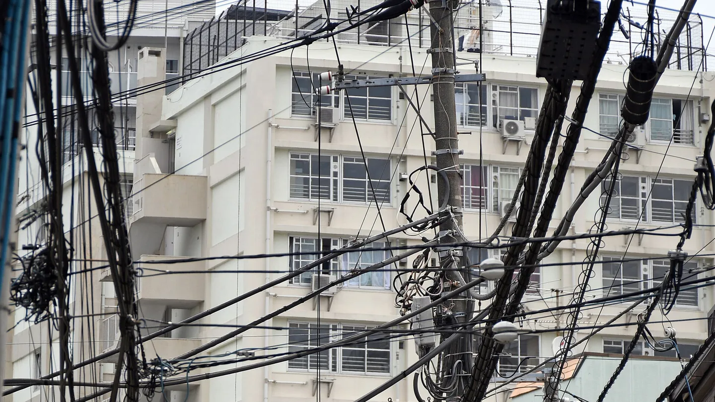 Dozens of black power and telecom cables strung between utility poles and tangled around transformers in front of a white apartment block in Takadanobaba, Tokyo
