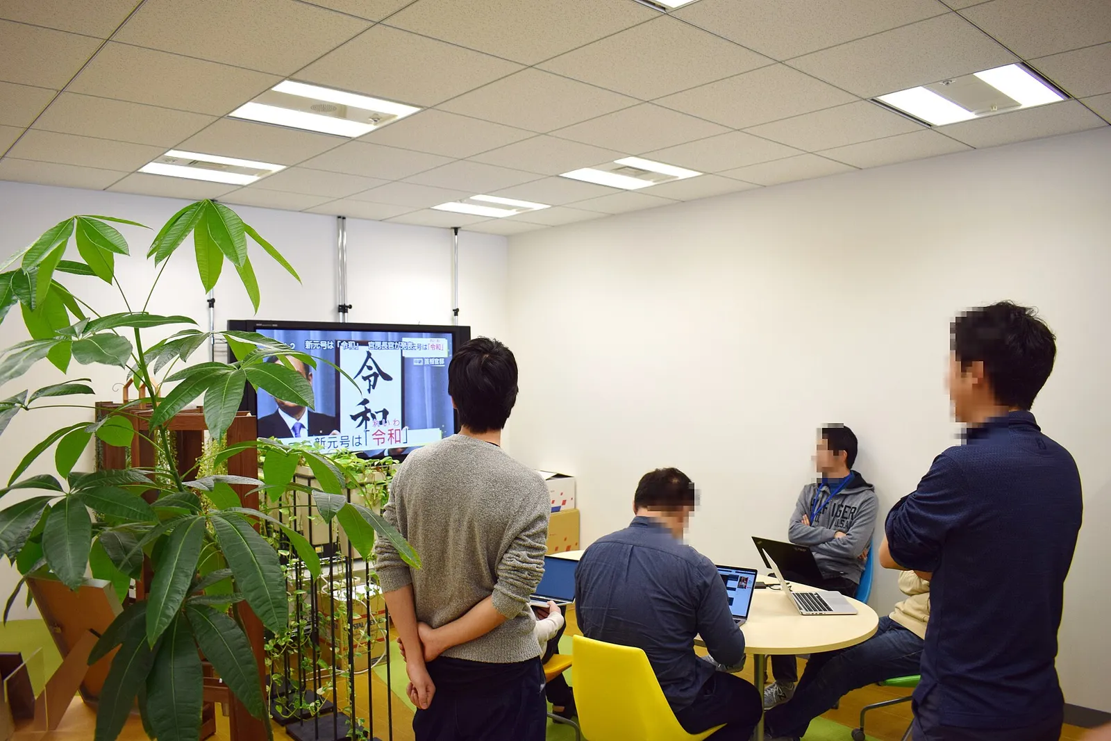 Four office workers in a Japanese office standing and sitting around a table facing a wall-mounted screen