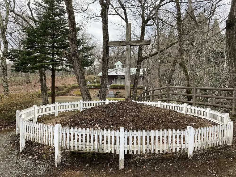 A grave mound inside a white picket fence, with a tall wooden cross behind it, in a wooded grove in Shingo