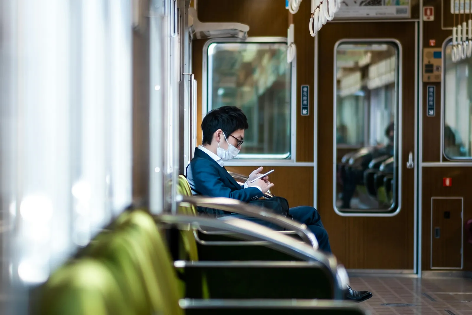 A man in a suit and face mask looking at his phone alone on an almost empty commuter train in Osaka