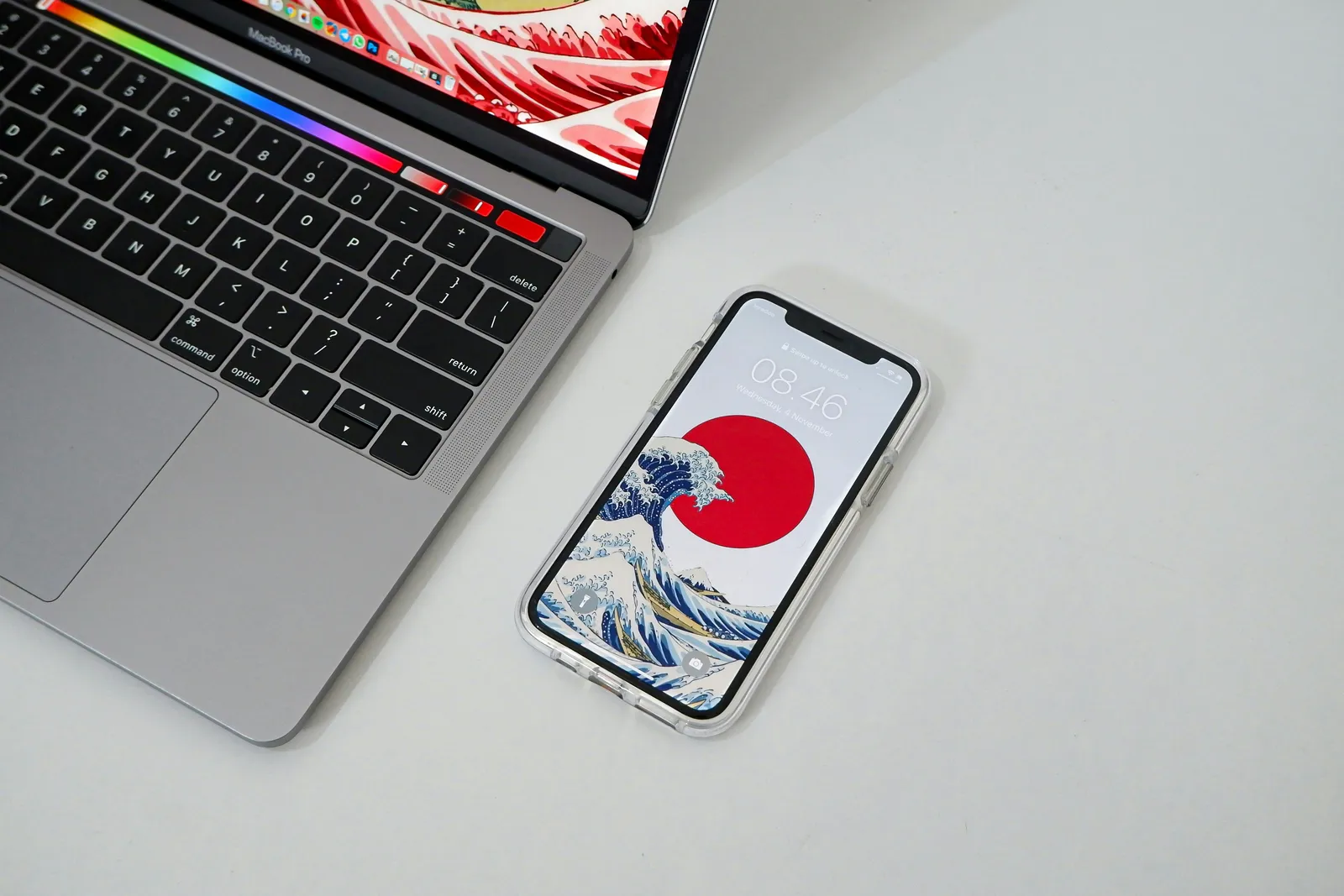 A MacBook Pro laptop and an iPhone with a Great Wave wallpaper lying side by side on a white desk