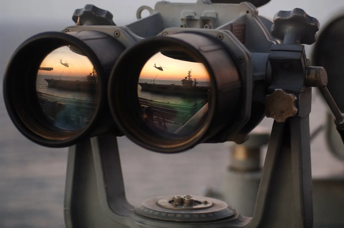 Large mounted binoculars on a ship's deck at sunset, with a helicopter and an aircraft carrier reflected in the lenses