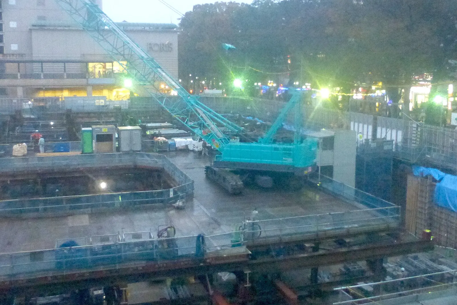 A teal crawler crane working on an excavated construction site in front of Fuchu Station at dusk
