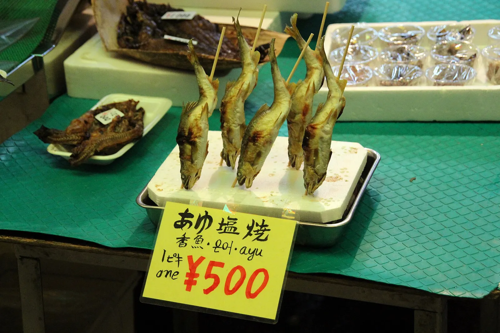 Grilled sweetfish on skewers at a Nishiki Market stall with a handwritten bilingual price sign reading 500 yen