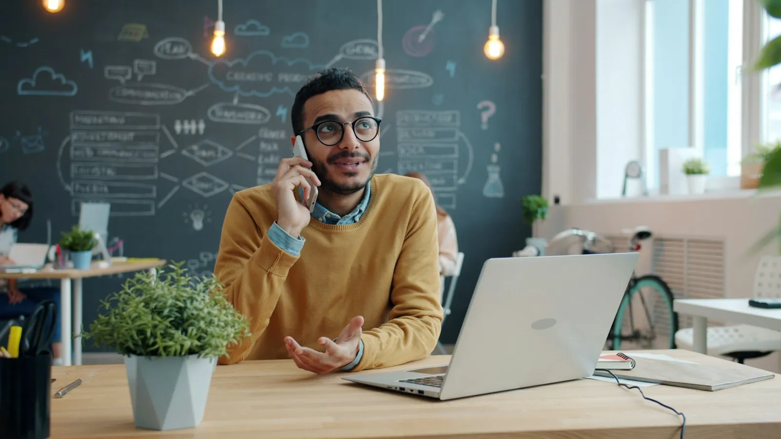 A man talking on a mobile phone at an office desk with an open laptop and a potted plant