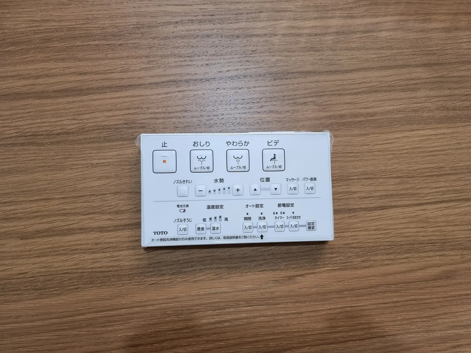 A white TOTO Washlet wall remote with Japanese button labels and small icons, lying on a wooden table