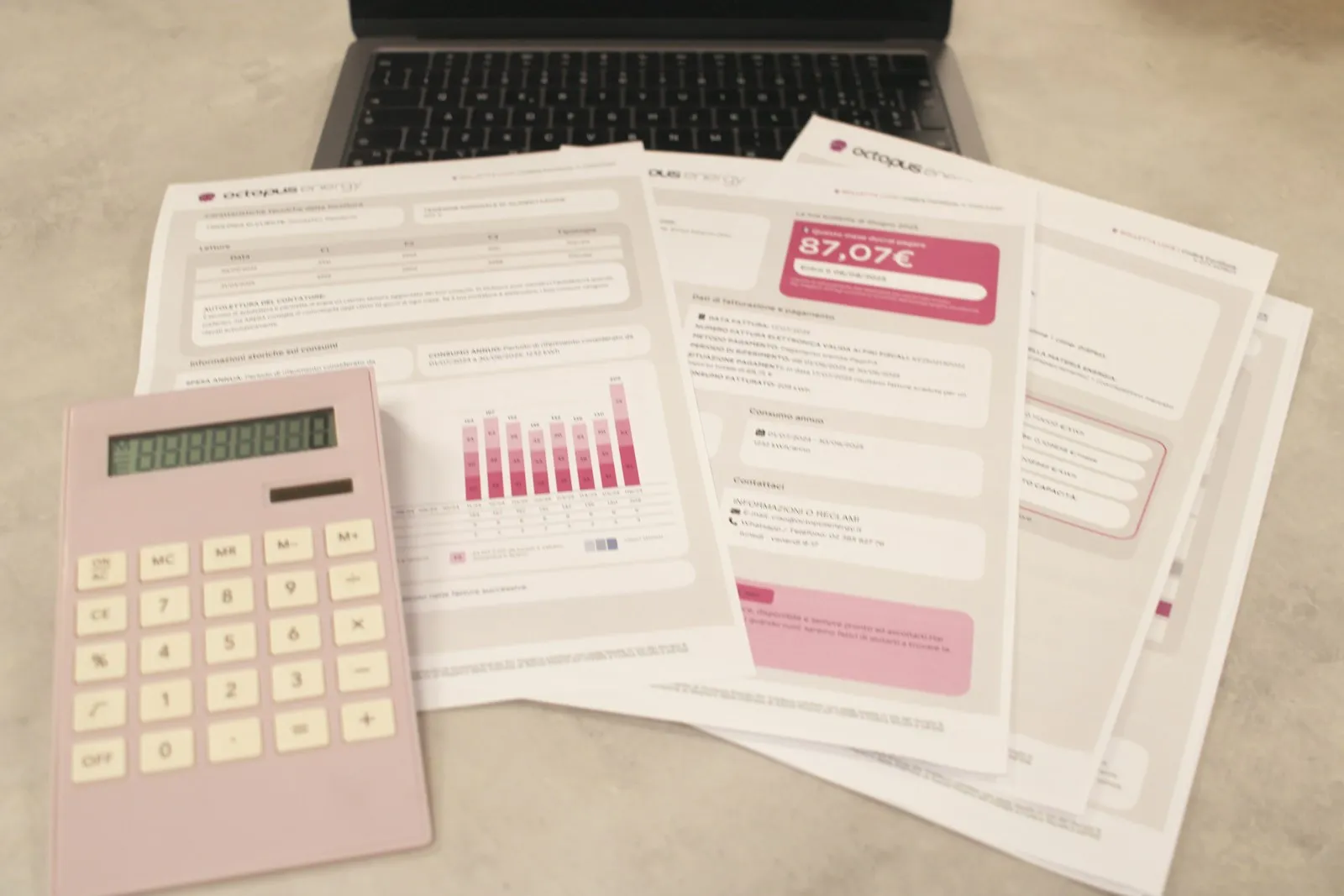 Printed bills and statements spread on a desk beside a pink calculator and an open laptop