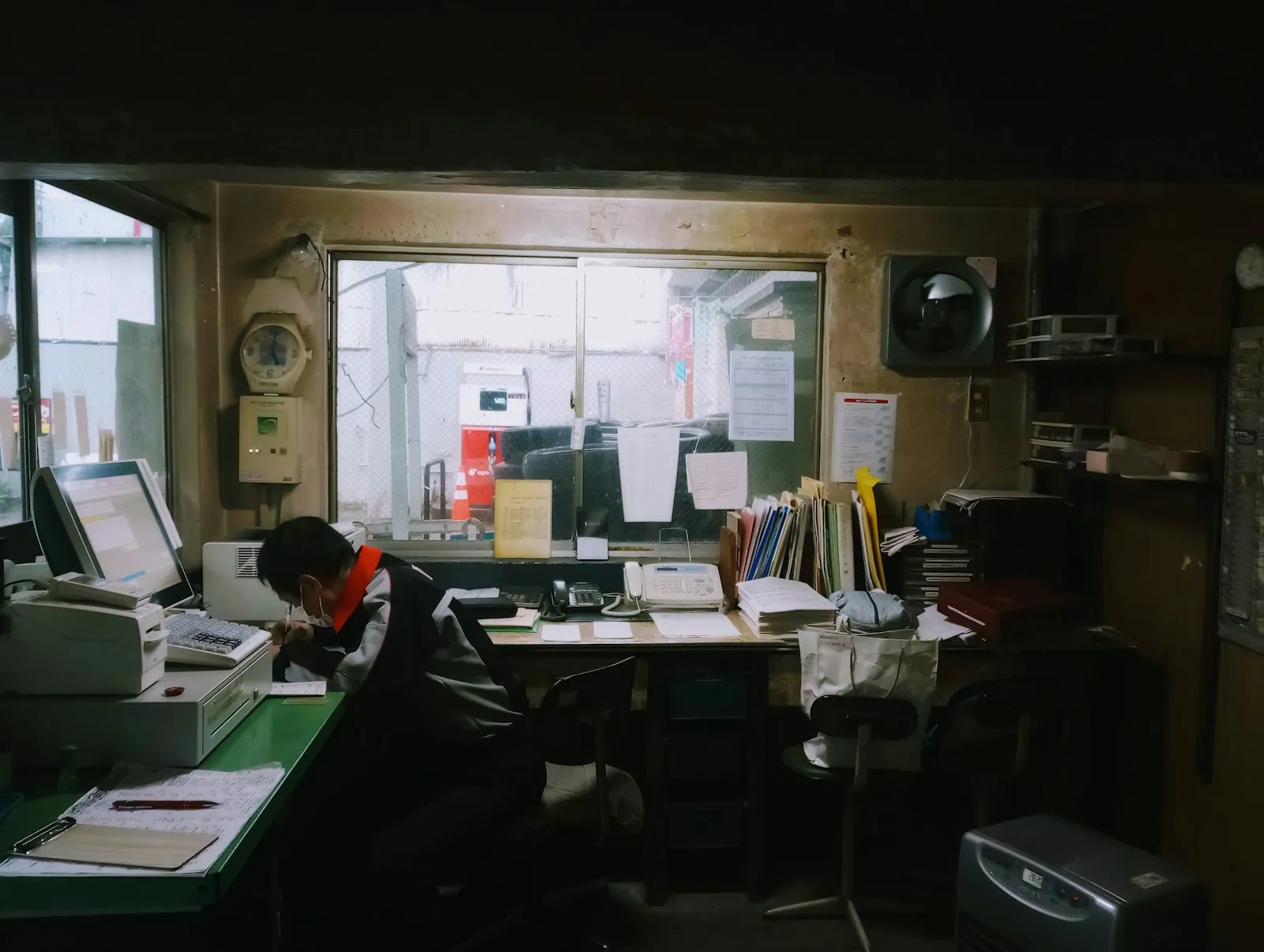 A man working at a cluttered desk in a small office with a computer, files and paperwork