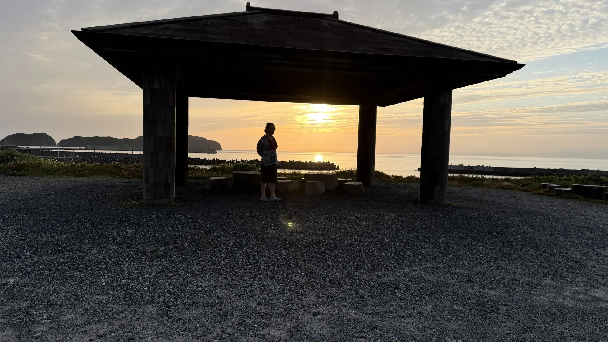 A person silhouetted under an open wooden pavilion by the sea as the sun sets over the water