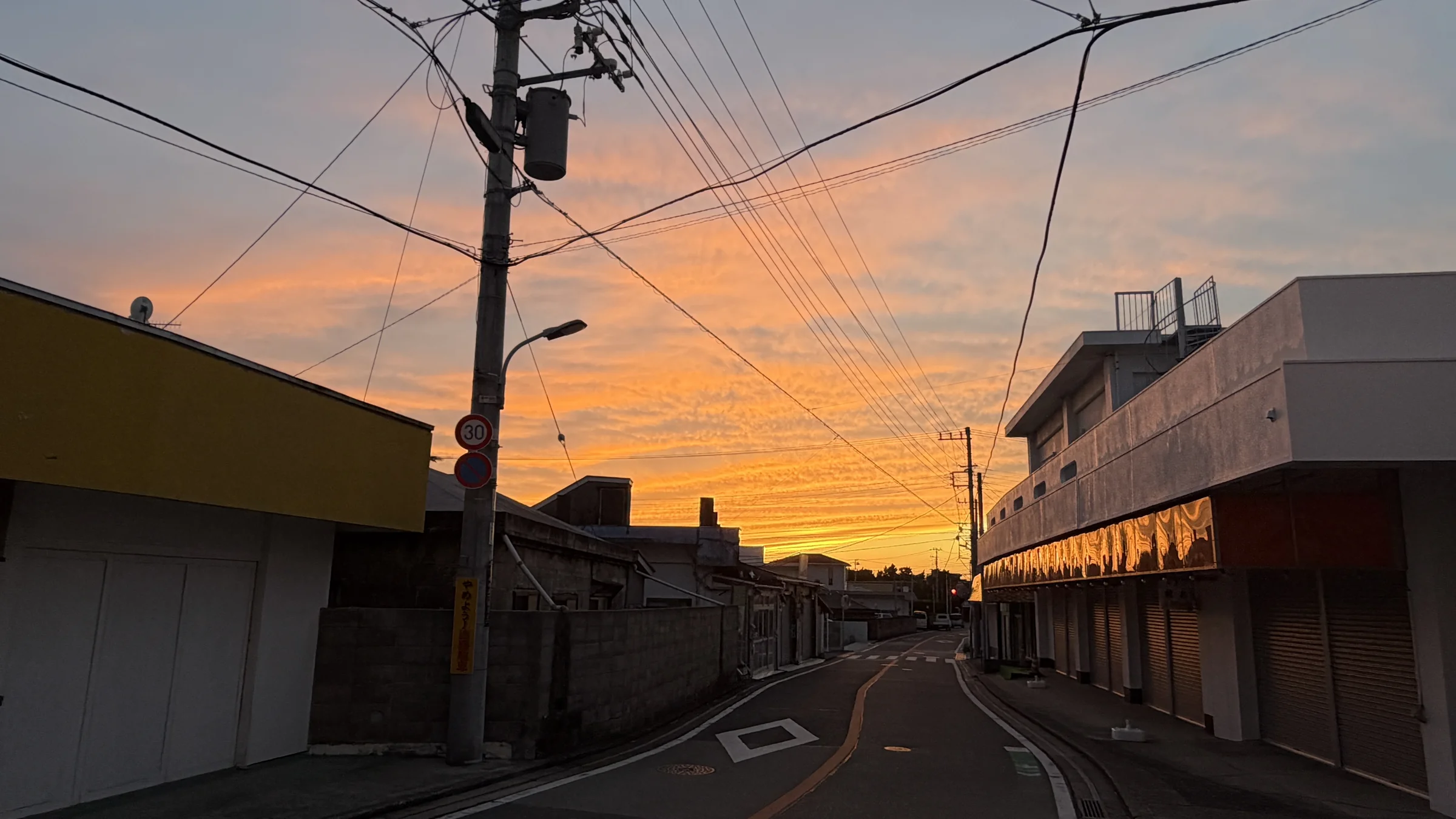 Quiet small-town street in Japan with closed shop shutters, utility poles and an orange evening sky